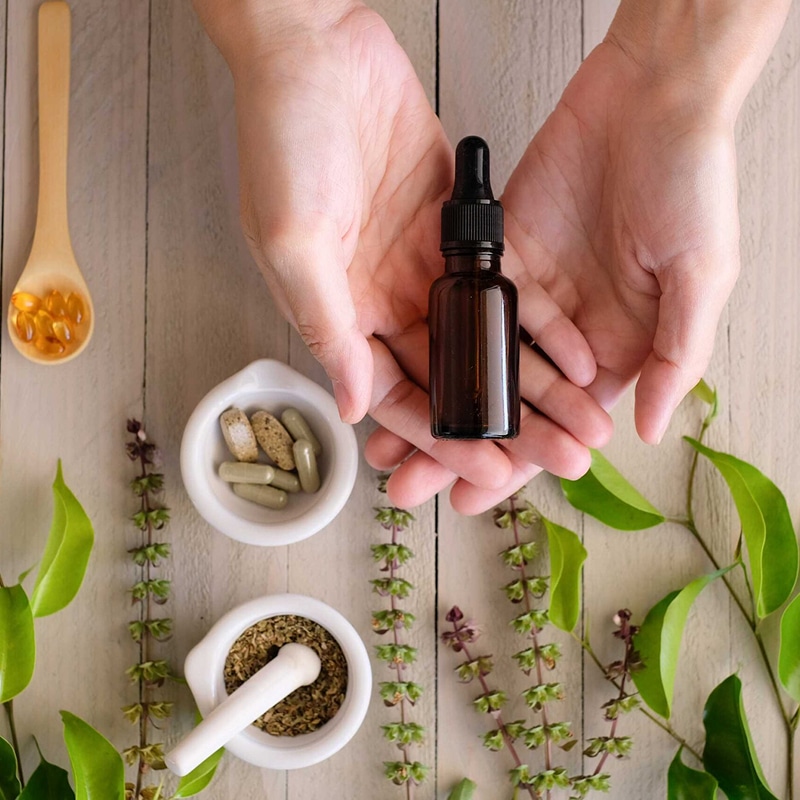 Mains tenant une fiole d'huile essentielle sur bois clair, entourée de plantes, gélules et mortier d'herbes médicinales.